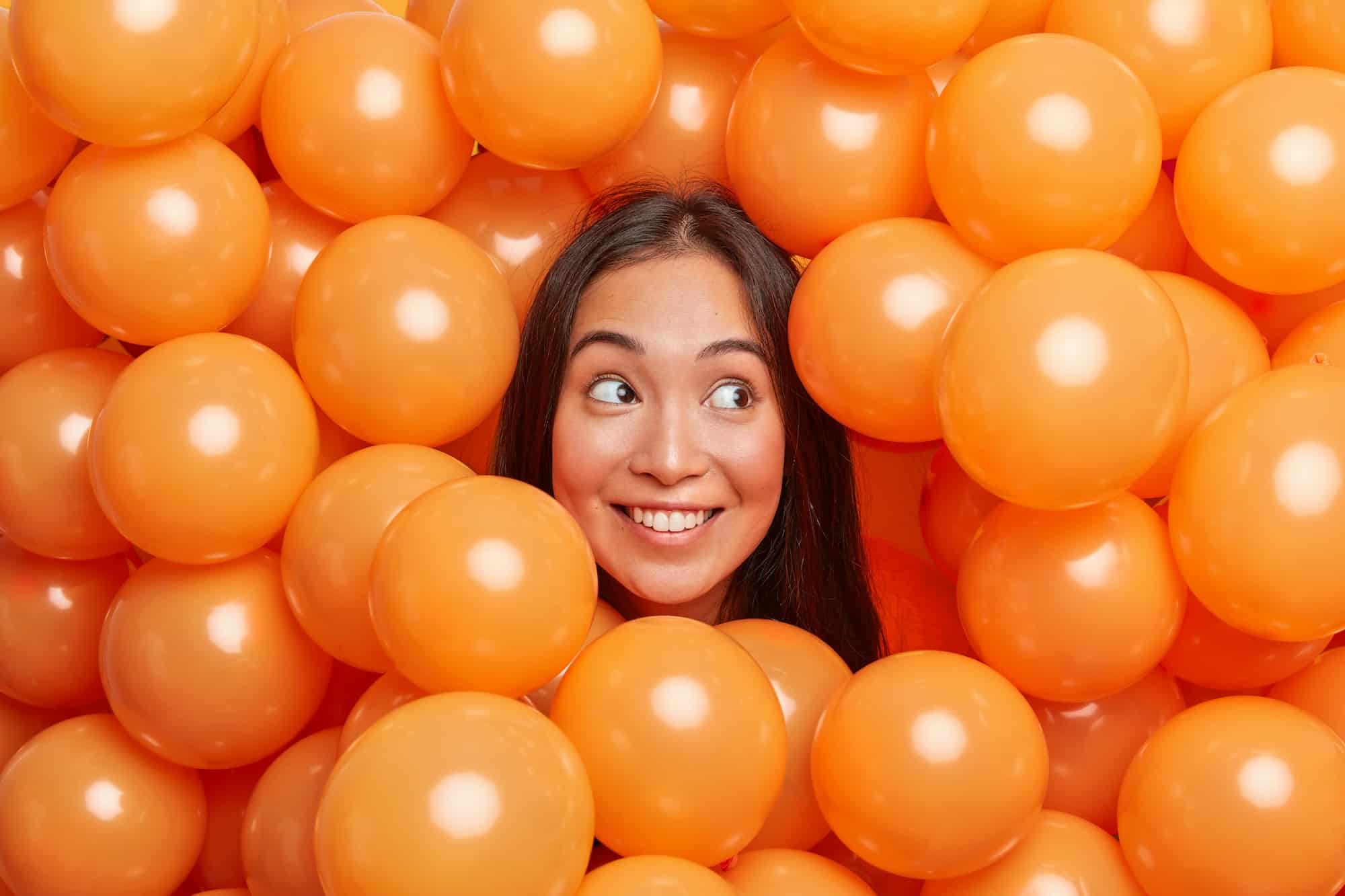 Positive young Asian woman smiles broadly and looks away surrounded by many orange air balloons enjo